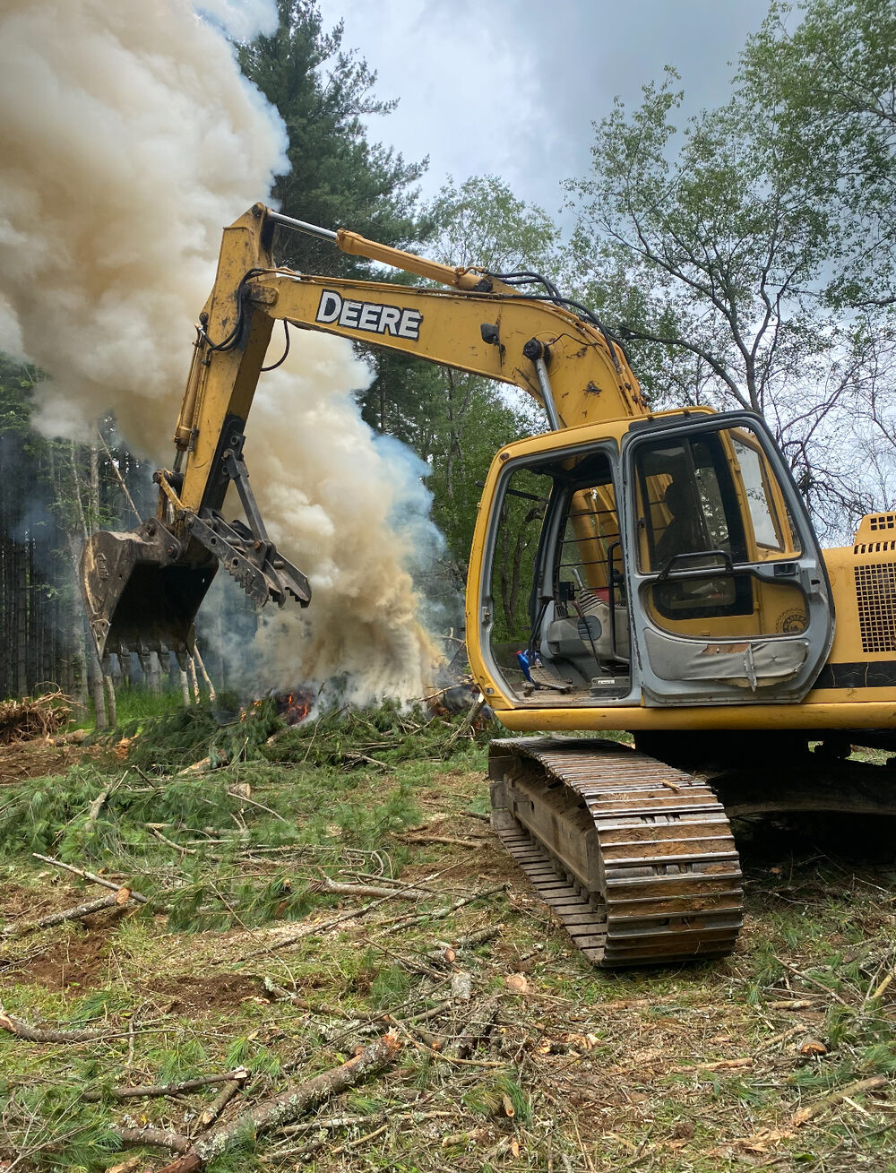 An excavator clearing land and throwing up a plume of dust on a wooded lot