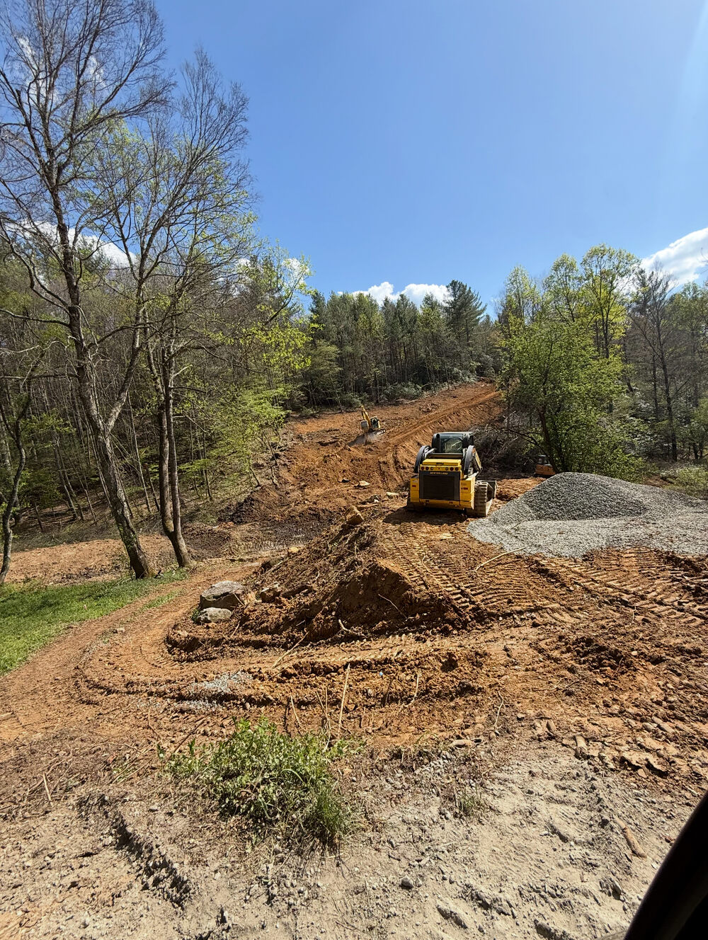A wide cleared and graded building site on a wooded mountain lot under a blue sky